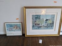 Two framed artworks leaning against a wall on floor; smaller framed print on left, larger framed lithograph on right