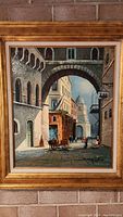 Full view of framed oil painting showing European street with archway, people, horse-drawn cart, and domed building in background.