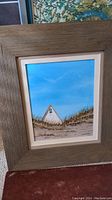 Close-up of small framed painting showing white house on sand dunes under blue sky with wooden frame and beige mat
