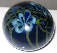 Top and angled views of spherical art glass paperweight with blue flowers and green tendrils against a deep blue background.