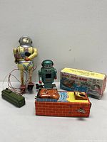 Three mechanical tin toys shown together: yellow spaceman robot, blue retro robot, and hippo bank with box.