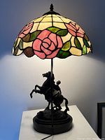 Lamp illuminated showing stained glass shade with rose motif and metal base of man riding rearing horse