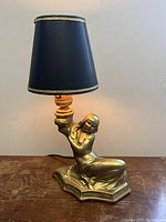 Full view of the bronzed metal Art Deco nude lady table lamp with black shade on wooden surface.