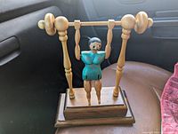 Front and side angled view of carved wooden mechanical gymnast man in teal outfit on a wooden high bar assembly, mounted on a rectangular base.