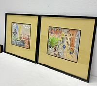 Two framed gouache paintings side by side showing Parisian street scenes with yellow matting and black frames.