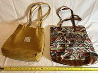 Two medium-sized used tote bags side by side on a white background with a measuring tape below them showing about 13 inches width each. Left bag is solid olive green with a white label, right bag has a southwestern multicolor pattern with brown trim and straps.