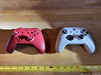Front view of two Xbox controllers on a wooden surface with measuring tape below