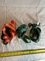 Photo of three used ratchet straps, two green and one orange, coiled with metal ratchets and hooks visible, lying on beige cloth with measuring tape below for scale.