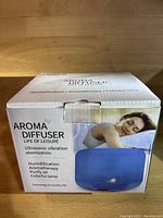 Closed retail box of the aroma diffuser showing product branding and features, including ultrasonic vibration atomization, humidification, aromatherapy, purification, and colorful lamp with photo of person resting next to diffuser.