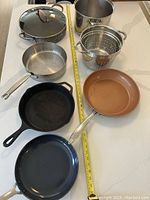 Photo of six assorted kitchen pots and pans arranged on floor with measuring tape for scale.