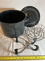 Black speckled enamelware pot with lid, metal rack, pot lifter placed in front with measuring tape showing size