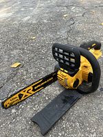 Photo of a yellow and black DeWalt cordless chainsaw with 12-inch blade labelled XR Brushless placed on asphalt ground with blade cover beside it.