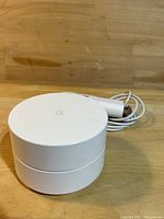 Photo of white Google WiFi Extender device and accompanying white power adapter coiled beside it on a wooden surface.