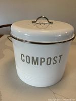 Front angle photo of closed white metal compost bucket with 'COMPOST' printed on the side and ventilated lid with metal handle.