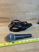 Photo of a black handheld microphone with a silver mesh grille, placed beside a measuring tape showing about 7 inches length, with a black cable coiled behind it.