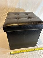 Front view of black padded storage ottoman showing button-tufted top and square shape with measuring tape at base