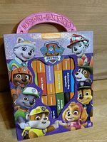 Closed view of Paw Patrol Book Block case showing 12 small block books inside with a purple background and Paw Patrol characters around the frame and a pink handle on top.