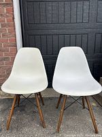 Two white molded plastic chairs with wooden legs and black metal rod supports placed outside against a garage door.
