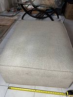 Top view of storage ottoman with light beige fabric and measuring tape for size reference