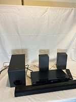 Sony surround sound system components arranged on a surface with a white backdrop: soundbar, subwoofer, two satellite speakers, DVD player, and remote control visible.