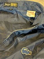 Close-up of the inside collar area showing size L label, material tag 100% nylon and a zipper with detail of the embroidered alligator logo patch.