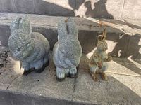 Three bunny statues arranged on outdoor stone stairs, two large gray concrete bunnies and one smaller rusted metal bunny.