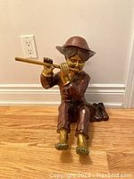Frontal view of bronze toned statue of boy sitting playing flute, wide-brimmed hat, bare feet visible, boots placed behind.