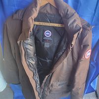 Full view of brown bomber style Canada Goose jacket hanging on a Burnett Limited wooden hanger against a blue backdrop showing interior padded lining and front zipper.