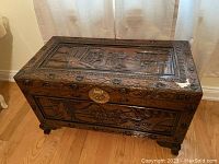 Full view of wooden chest showing carved top panel and metal lock, wood floor background