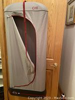 Full view of the CHI portable clothing steamer with beige fabric enclosure and maroon trim, zipper partially open revealing interior hanger.