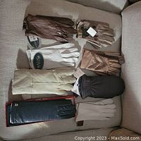 Seven pairs of assorted ladies gloves laid on a couch, various shades of brown, beige, black and white, some with tags and boxed packaging.