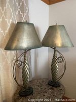 Pair of tall pewter tone table lamps shown side by side on a table against a wall with patterned curtain.