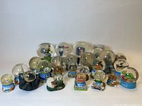 Wide view of 24 assorted snow globes from various international locations showing different figurines, city and animal themes.