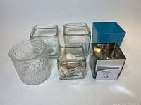 Image showing six vases: one clear textured round vase, three clear glass cube vases, blue square vase, silver reflective square vase; one glass vase contains seashells.