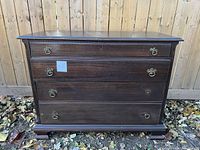 Front view of antique wood dresser showing four drawers and brass pull handles, set against a wooden fence outdoors with fallen leaves.