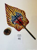Balinese double sided painted fan with a folk art style face, along with a round wooden stand with floral design, placed on white background with lot number card.
