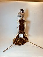 Full front view of Balinese puppet sculpture with wooden sticks crossed at base wrapped in patterned fabric.