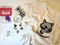 Overview of all items in the lot including T-shirt, plate, pins, glass cat, and mochi squish toys