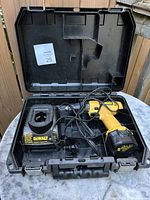 Black hard plastic carrying case open showing yellow and black DeWalt cordless drill with attached battery and black DeWalt battery charger with power cord inside.