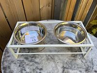 Top side angle showing two stainless steel dog bowls set inside a white metal frame holder on a concrete surface outdoors.