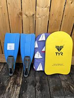 Blue swim flippers next to two swim flutter boards, one yellow with TYR logo and one with sailboat pattern, set against a wood background.