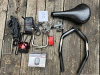 Top-down photo of various black bicycle parts laid out on a wooden surface showing saddle, handlebars, pedals, inner tubes, lock, bottle cage, rear light, and packaged bike computer.