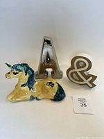 Three decorative items: yellow and blue ceramic unicorn figurine, silver light-up letter 'A', and ceramic ampersand symbol with wood texture.