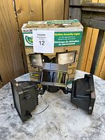 Outdoor motion sensor security light with dual adjustable floodlights and central sensor, with original box in background.