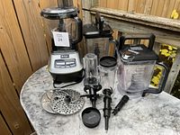 Ninja food processor with multiple accessories including blender container, blades, and small blending cup, displayed on a small round table outside.