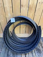 Photo shows a tightly coiled roll of black ribbed plastic garden edging against a wooden fence background.