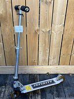 Silver aluminum kick scooter with black foam handlebar grips and black deck showing Infinity branding, leaning against wooden fence on wooden deck.