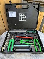 Open black plastic tool case showing green handled pliers, hammer, wrench, screwdriver, and Allen keys set in molded slots with some missing tools.