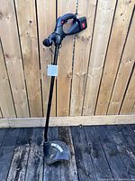 Full view of black electric weed whacker standing upright with power cord and trigger handle visible against wooden fence background.