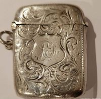 Front view of silver match safe showing shield-shaped monogram and floral scroll engravings